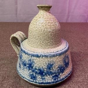 May include: A handmade ceramic pitcher with a lid. The pitcher has a light beige, textured body with a blue and white speckled design around the base. It features a curved handle and a small, rounded lid.