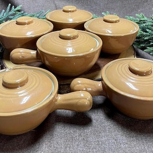 May include: A set of six tan ceramic soup bowls with matching lids. Each bowl has a handle and a circular lid with a knob. The bowls are arranged on a wooden surface with greenery in the background.