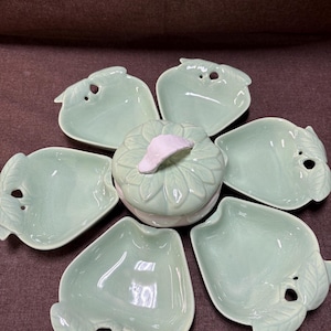 May include: A light green ceramic serving set. The set includes six leaf-shaped dishes and a round lidded container with a leaf-like design. The dishes have a small hole and a leaf-shaped accent. The container has a white accent on the lid.