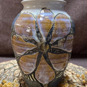 May include: A ceramic vase with a brown, purple, and white glaze. The vase is decorated with a large, embossed flower design.