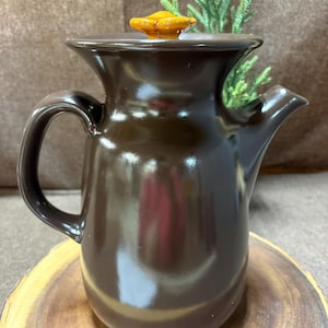May include: A brown ceramic teapot with a handle and spout, featuring a lid with an orange knob. The teapot is placed on a wooden surface, showcasing its glossy finish and classic design. The teapot is a kitchenware item.