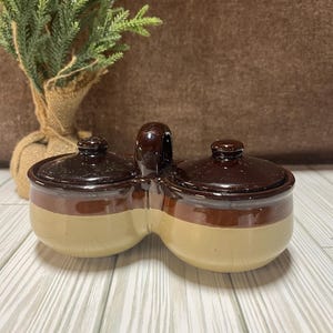 May include: A brown and beige ceramic double crock with a handle. The crock has two separate compartments with lids.