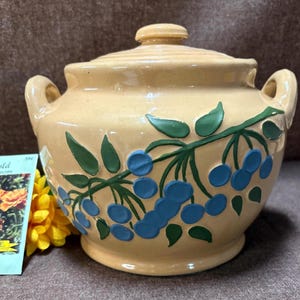 May include: A cream-coloured ceramic lidded pot with two handles and a decorative branch design featuring blue berries and green leaves. A yellow flower and a seed packet are next to the pot.