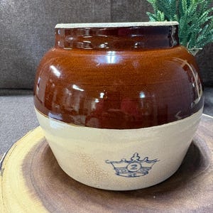 May include: A vintage brown and cream-colored stoneware crock. The upper portion is a glossy brown, while the lower half is cream with a blue crown and the number 2. The crock is sitting on a wooden slice.