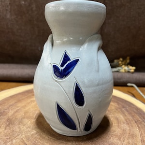 May include: A small, white ceramic vase with a blue floral design. The vase has a rounded body and a narrow neck.