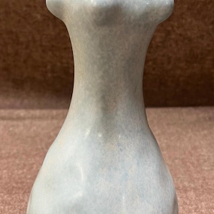 May include: A light blue ceramic vase with a unique, abstract, human-like form. The vase has a textured surface and a rounded base.