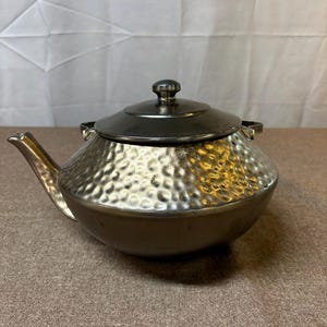 May include: A metallic teapot with a hammered texture on the upper body and a smooth, dark finish on the lower section. It features a curved spout, two small side handles, and a removable lid with a central knob, all in a polished silver tone.