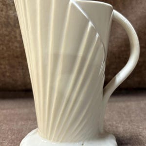 May include: Cream-colored ceramic pitcher with a unique, fluted design. The pitcher features a handle and a wide, flared top. The base is a rounded, scalloped shape. The overall design is reminiscent of a seashell.
