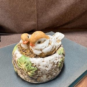May include: A ceramic bird figurine featuring two birds nestled in a detailed nest. The birds have brown and white feathers and orange beaks. The nest is a light beige with green leaf accents, creating a charming decorative piece.