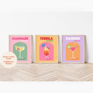 May include: Three colorful posters with illustrations of cocktails. The posters are framed in wood and have a pink, yellow, and purple background. The text on the posters reads "Champagne Cocktail", "Tequila Sunrise", and "Daiquiri Dream".