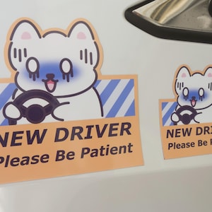 May include: Two rectangular stickers featuring a cartoon white dog with blue shading around its eyes, holding a black steering wheel. The background has blue diagonal stripes and a peach border. The text reads NEW DRIVER Please Be Patient.