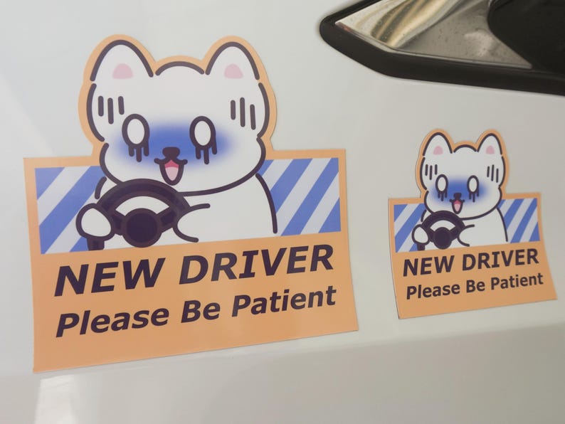 May include: Two rectangular stickers featuring a cartoon white dog with blue shading around its eyes, holding a black steering wheel. The background has blue diagonal stripes and a peach border. The text reads NEW DRIVER Please Be Patient.