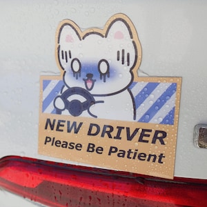 May include: A rectangular car decal featuring a cartoon white cat with blue shading on its face, holding a black steering wheel. The background has blue and white diagonal stripes. The text reads NEW DRIVER Please Be Patient in black font.