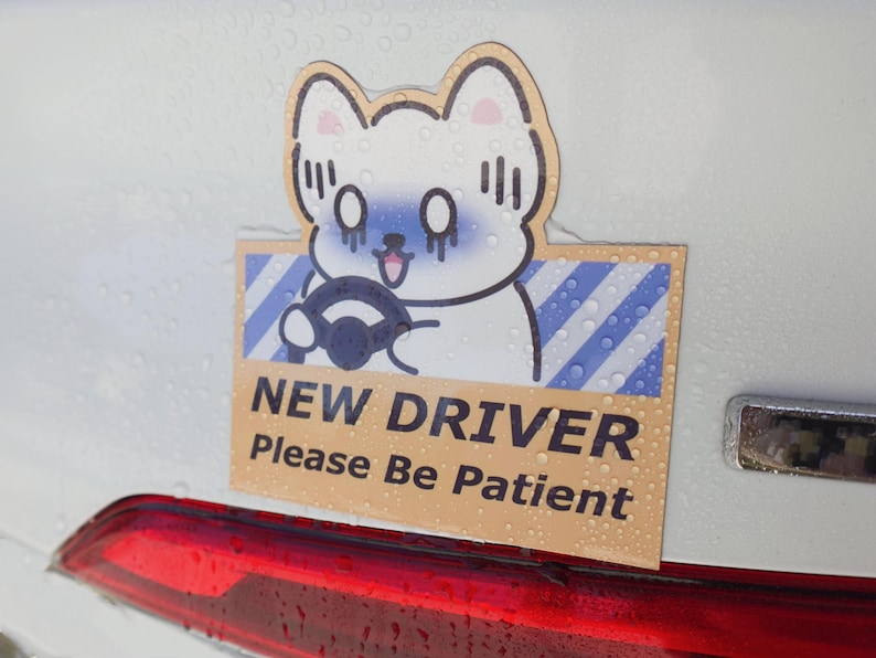 May include: A rectangular car decal featuring a cartoon white cat with blue shading on its face, holding a black steering wheel. The background has blue and white diagonal stripes. The text reads NEW DRIVER Please Be Patient in black font.
