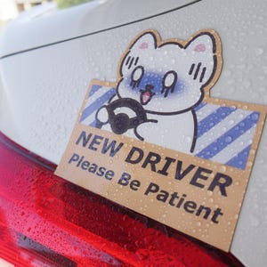 May include: A rectangular car decal featuring a cartoon white cat with a blue face driving a vehicle. The background has blue diagonal stripes, and the text reads NEW DRIVER Please Be Patient in bold black letters on a tan background.