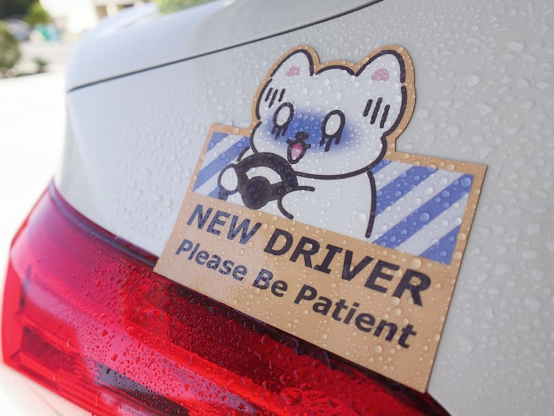 May include: A rectangular car decal featuring a cartoon white cat with a blue face driving a vehicle. The background has blue diagonal stripes, and the text reads NEW DRIVER Please Be Patient in bold black letters on a tan background.