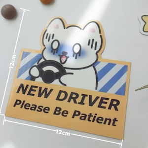 May include: A square sticker featuring a white cartoon cat driving a car with blue diagonal stripes in the background. The text reads NEW DRIVER Please Be Patient in black. A white ruler overlay indicates the sticker measures 4.72 inches by 4.72 inches.