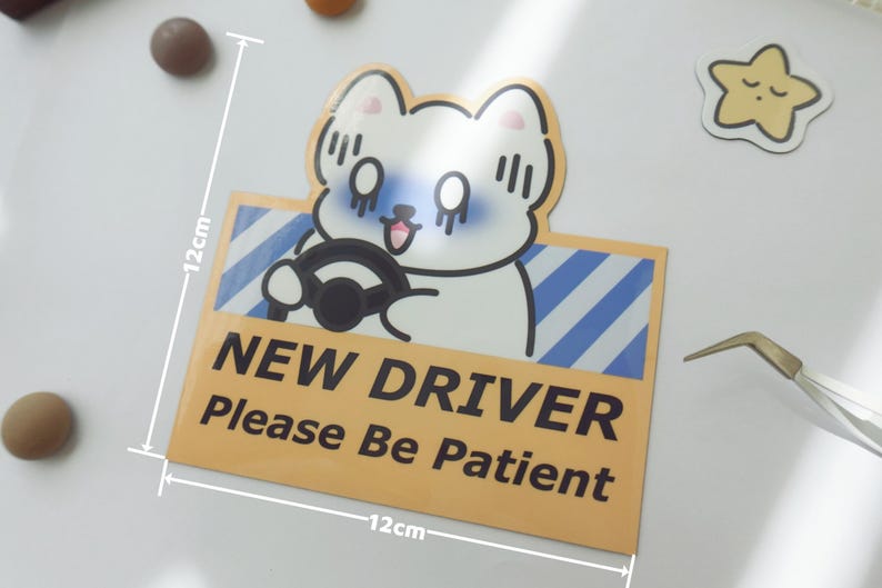 May include: A square sticker featuring a white cartoon cat driving a car with blue diagonal stripes in the background. The text reads NEW DRIVER Please Be Patient in black. A white ruler overlay indicates the sticker measures 4.72 inches by 4.72 inches.