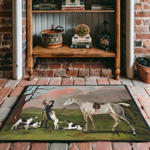 May include: A vintage-style doormat featuring a painting of a man on horseback with three dogs. The painting is set in a field with trees and a pink sky in the background.