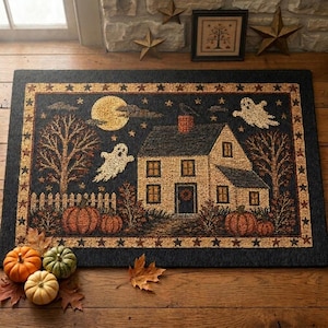 May include: A rectangular rug featuring a dark background with a central house, two white ghosts, a yellow moon, and orange pumpkins. The border is decorated with a repeating star pattern. The rug is placed on a wooden floor with three real pumpkins.