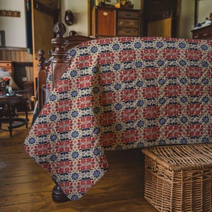 May include: A floral patterned quilt in red, blue, and cream covers a wooden bed frame. The quilt features a repeating pattern of stylized flowers and leaves. The bed is in a room with wooden floors and furniture, and a wicker basket is at the foot of the bed.