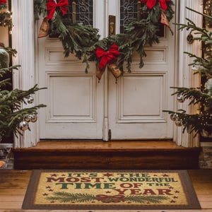 May include: A rectangular doormat with the text "IT'S THE MOST WONDERFUL TIME OF THE YEAR" in red and green, surrounded by stars and a decorative border. The mat is placed in front of a white door decorated with a Christmas wreath and red bows.