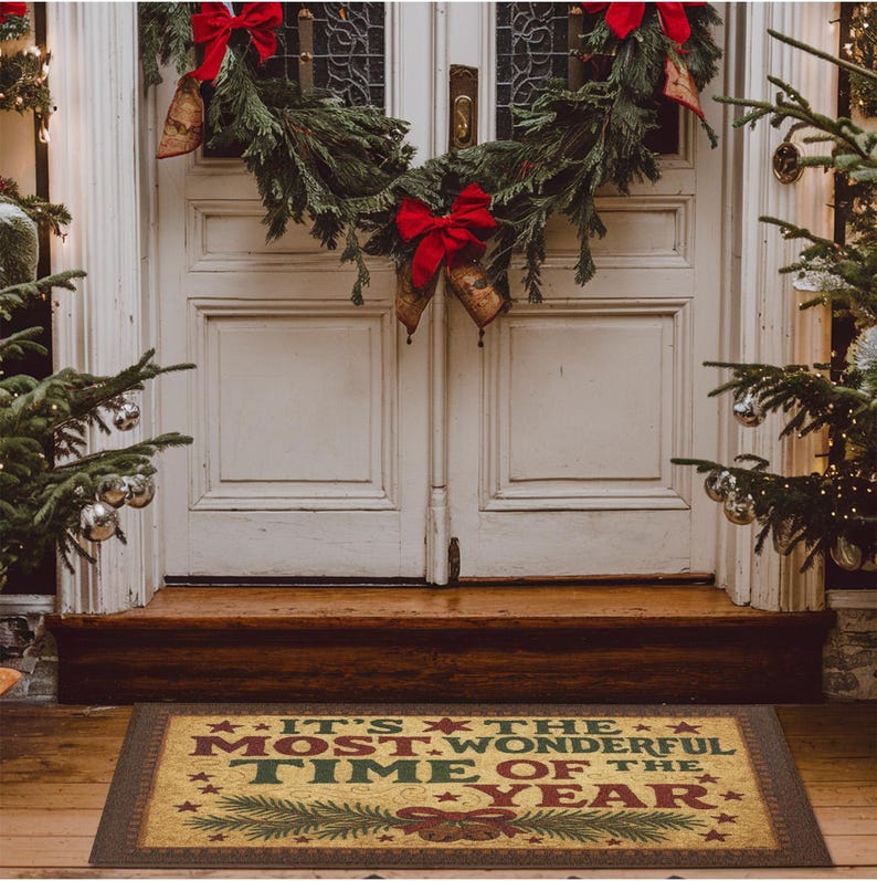 May include: A rectangular doormat with the text "IT'S THE MOST WONDERFUL TIME OF THE YEAR" in red and green, surrounded by stars and a decorative border. The mat is placed in front of a white door decorated with a Christmas wreath and red bows.