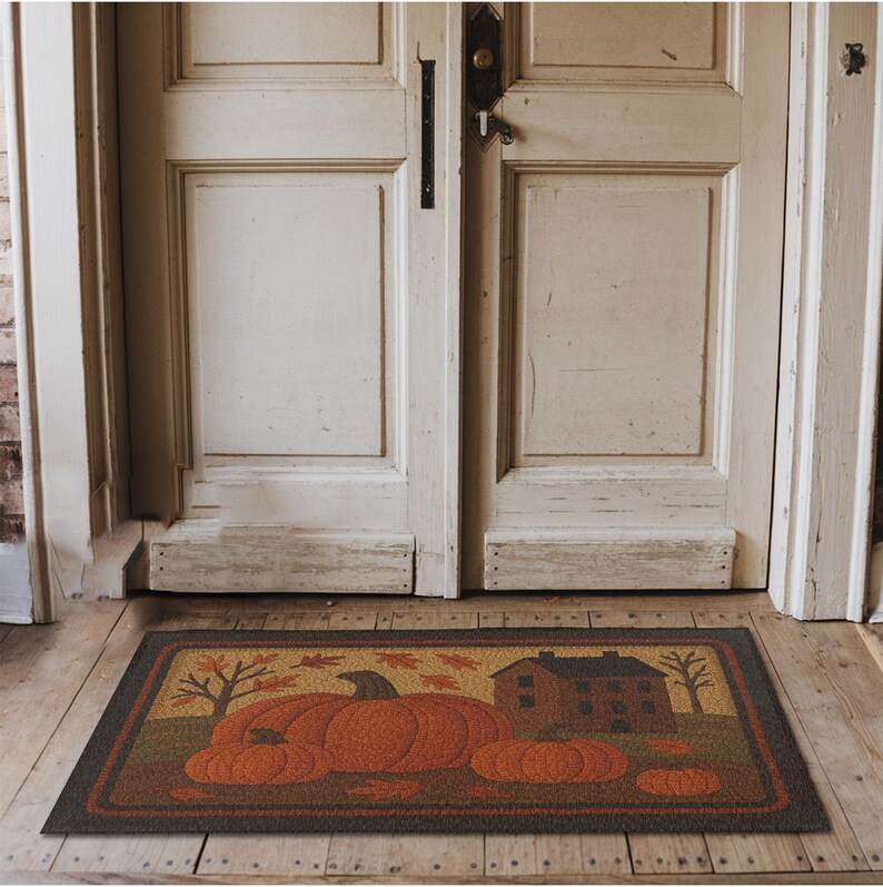 May include: Rectangular doormat featuring a fall harvest scene. The mat displays three large orange pumpkins, a small house, and trees with falling leaves. The border is dark brown, and the background is a mix of brown and tan.