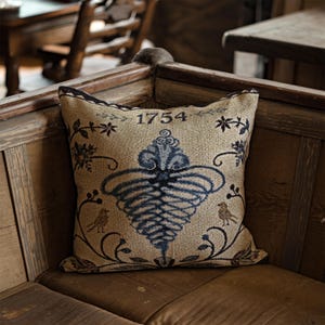 May include: Square decorative pillow with a beige background and a blue and black floral design. The pillow features the year "1754" and a central stylized design with birds and floral accents. The pillow is placed on a wooden bench.