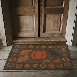 May include: A rectangular doormat with a brown background and a fall-themed design. The design includes a large orange pumpkin, oak leaves, a black crow, and decorative circles in shades of orange and yellow. The mat is placed in front of a wooden door.