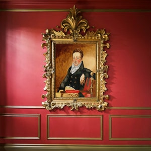 May include: A framed portrait of a man in a dark suit and white cravat, sitting in a chair with a book in his lap. The frame is ornate and gold, and the background is a red wall.
