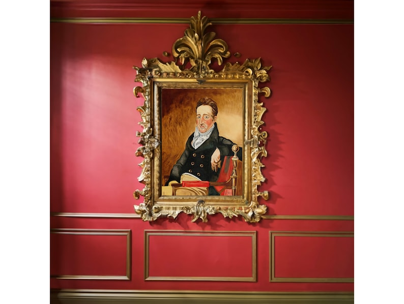 May include: A framed portrait of a man in a dark suit and white cravat, sitting in a chair with a book in his lap. The frame is ornate and gold, and the background is a red wall.