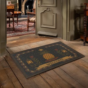 May include: Rectangular doormat with a dark blue background and a pineapple design. The mat features a gold banner with the word "WELCOME" and decorative floral and lantern motifs. The mat is placed on a wooden floor.