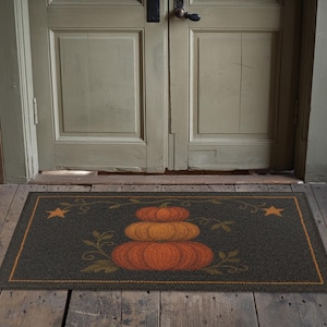 May include: A rectangular doormat with a dark grey background and a border. The mat features a stack of orange pumpkins with green leaves and vines, and two yellow stars. The mat is placed in front of a green door.