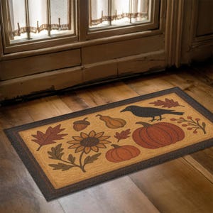 Può includere: Zerbino rettangolare con bordo marrone e centro beige con illustrazioni a tema autunnale. Il design include un corvo nero, zucche, foglie, un girasole e una ghianda. Il tappeto è posto davanti a una porta.