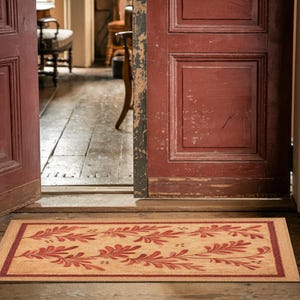 May include: A rectangular doormat with a beige background and a red floral design. The border is a darker shade of red. The mat is placed in front of a red door, creating a welcoming entrance.