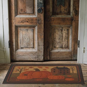 May include: Rectangular doormat featuring a fall harvest scene. The mat displays a large orange pumpkin, smaller pumpkins, a brown house, and trees with falling leaves. The border is a dark brown color. The mat is placed in front of a rustic wooden door.