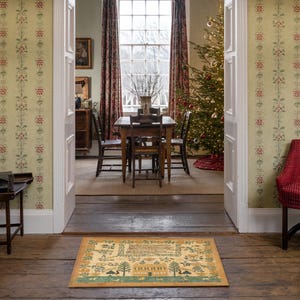 May include: A brown and beige patterned doormat with a Christmas tree design and the text "O Come, O Come Emmanuel" in a green font. The doormat is placed in front of a doorway with a wooden floor and a Christmas tree in the background.