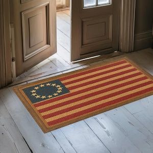 May include: Rectangular doormat featuring a colonial American flag design. The mat has red and yellow stripes with a blue square in the upper left corner containing a circle of yellow stars. The mat is framed with a brown border.