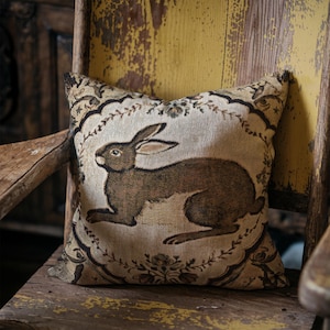 Puede incluir: Cojín cuadrado con una ilustración de conejo marrón sobre fondo crema, enmarcado por un borde floral y de volutas. El cojín está colocado sobre una silla de madera envejecida, añadiendo un toque rústico a la decoración.