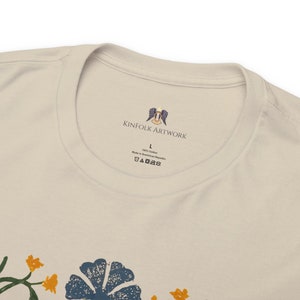 May include: A white t-shirt with a floral print and a label that says "Kinfolk Artwork" with a logo of a bird with wings spread. The label also says "100% Cotton" and "Made in Dominican Republic".
