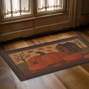 May include: Rectangular doormat with a fall theme. The mat features a large orange pumpkin, a house, and autumn leaves. The border is dark brown, and the background is tan and brown.