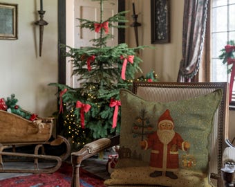 May include: A festive Christmas scene featuring a decorated evergreen tree with red bows and lights. A decorative pillow with a Santa Claus design sits on a striped chair. A wooden sleigh and red rug add to the holiday ambiance.