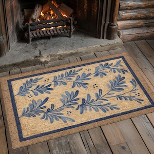 May include: Rectangular doormat with a natural tan background and a navy blue border. The mat features a repeating pattern of blue leaves and stems. The mat is made of a woven material and is placed in front of a fireplace.