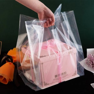 May include: A clear plastic bag with handles, containing a pink gift box with a white ribbon and the words "Happy Birthday" printed on it.