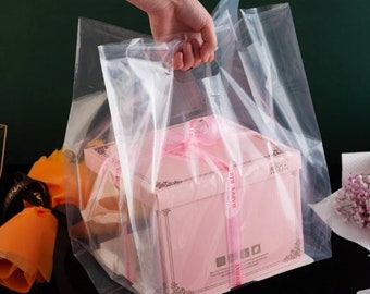 Puede incluir: Una bolsa de plástico transparente con asas, que contiene una caja de regalo rosa con una cinta blanca y las palabras "Feliz cumpleaños" impresas en ella.