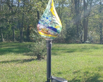 May include: A colorful, teardrop-shaped glass garden stake with a solar panel. The glass art features swirls of yellow, blue, green, and pink. The stake is mounted on a black metal pole, perfect for outdoor decoration in a garden or yard.