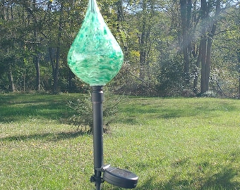 May include: A green glass garden stake with a teardrop-shaped top. The glass has a marbled appearance with shades of green and white. The stake is mounted on a black metal pole with a solar panel attached, set in a grassy yard.