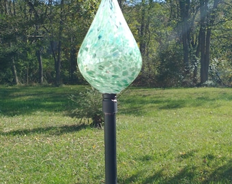 May include: A decorative outdoor solar light with a teardrop-shaped glass shade. The shade is a light green color with white speckles. The light is mounted on a black metal pole and has a solar panel at the base.