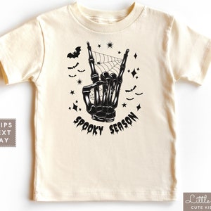 May include: A cream colored t-shirt with a black skeleton hand making a rock and roll sign. The hand is surrounded by bats, stars, and a spider web. The text "Spooky Season" is written below the hand.